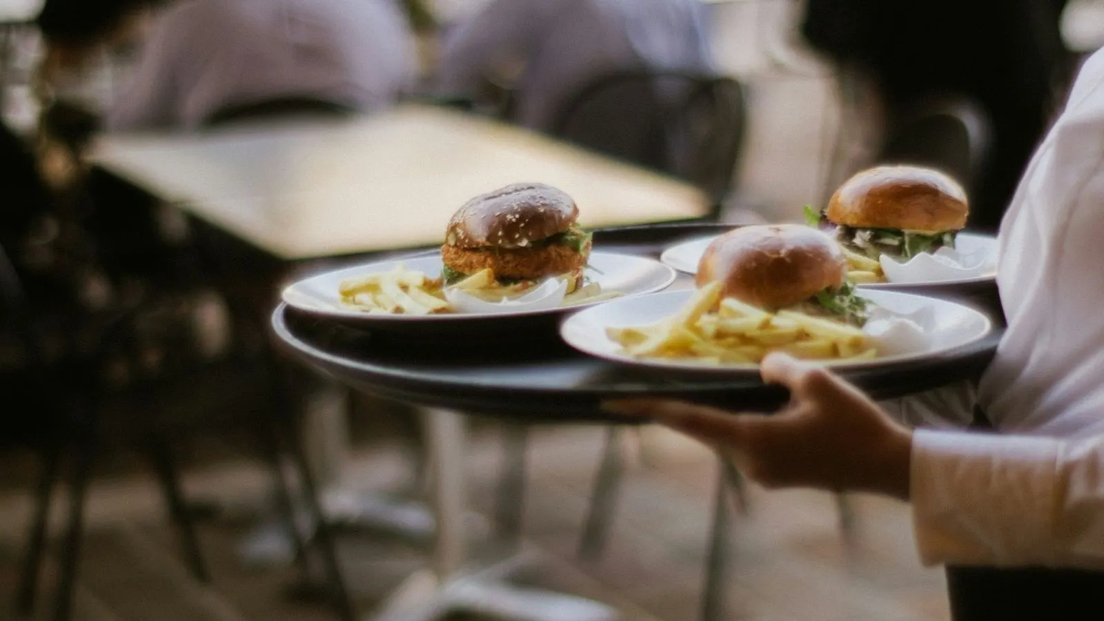 Kamarier duke çuar tri pjata me burger në një tarracë plot klientë